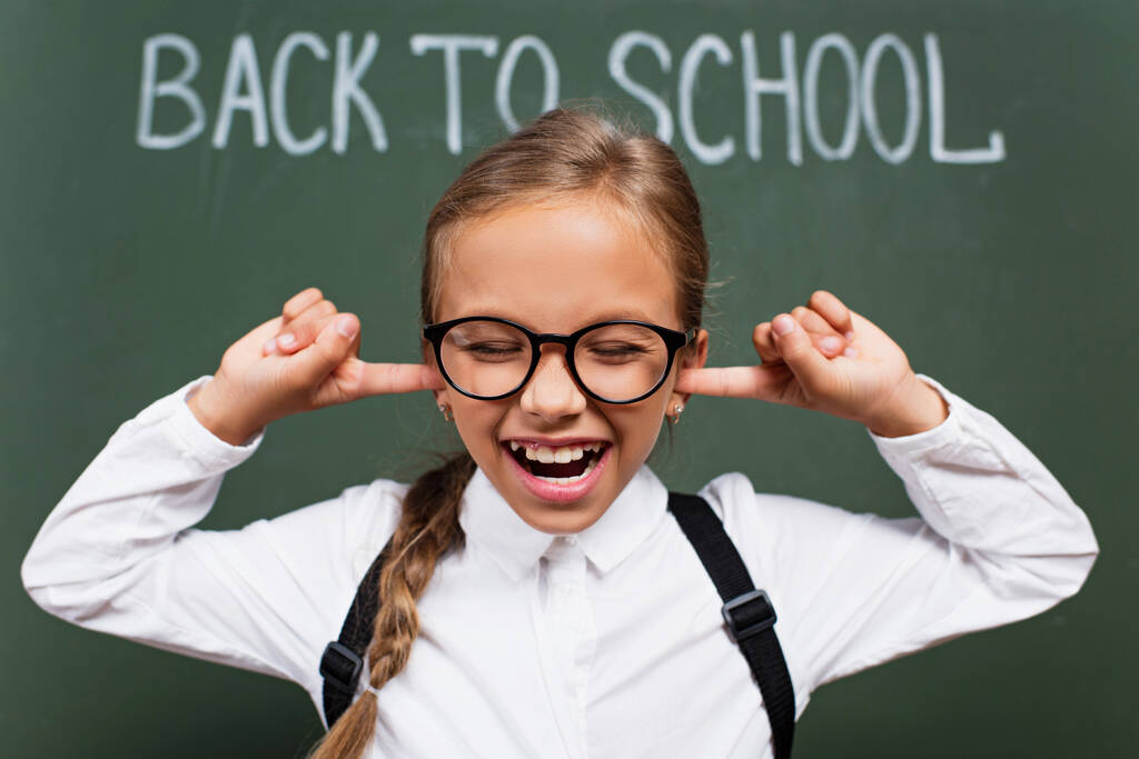 selective focus of displeased schoolgirl in eyeglasses plugging ears with fingers near back to school inscription on chalkboard - Photo, Image