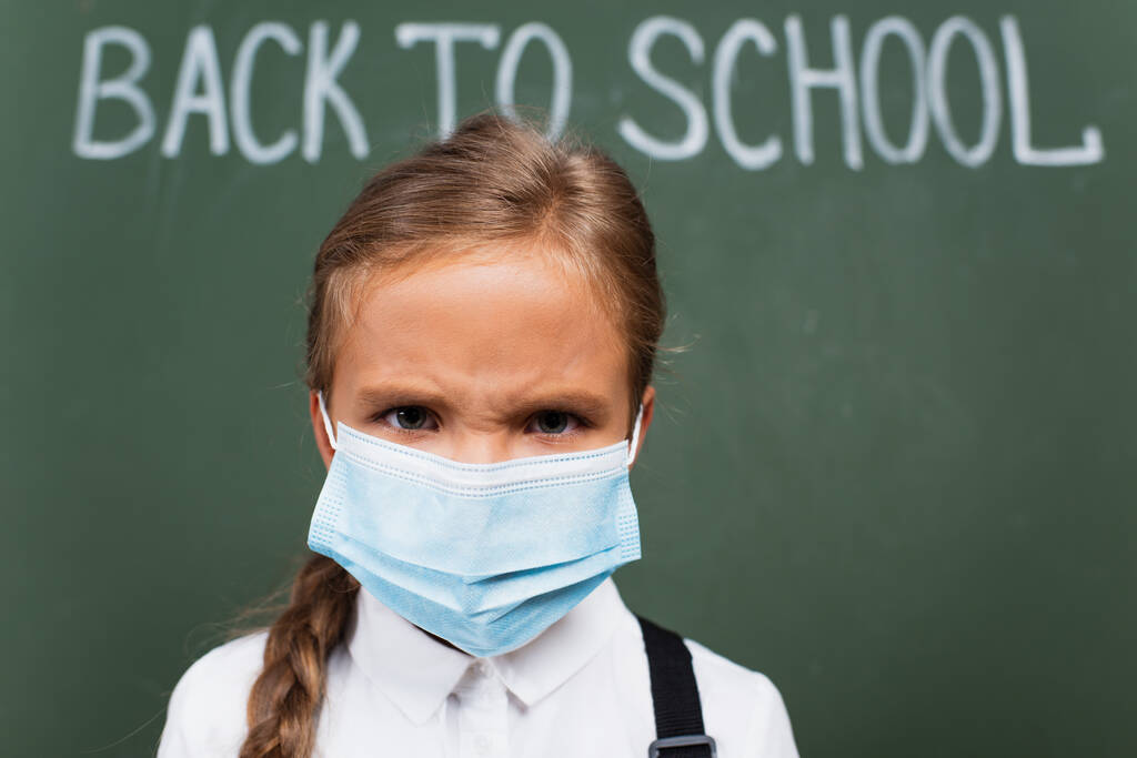 επιλεκτική εστίαση του κατσούφιασμα μαθήτρια σε ιατρική μάσκα κοιτάζοντας κάμερα κοντά στο chalkboard με πίσω στο σχολείο επιγραφή - Φωτογραφία, εικόνα
