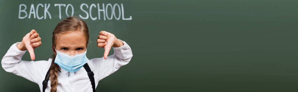 πανοραμικός προσανατολισμός δυσαρεστημένης μαθήτριας με ιατρική μάσκα που δείχνει τους αντίχειρες προς τα κάτω κοντά στο chalkboard με γράμματα πίσω στο σχολείο - Φωτογραφία, εικόνα