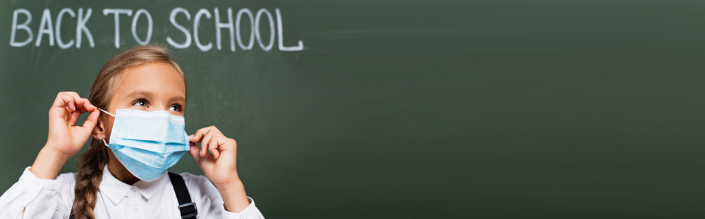 панорамная концепция школьницы, надевающей медицинскую маску рядом со школьной надписью на доске - Фото, изображение