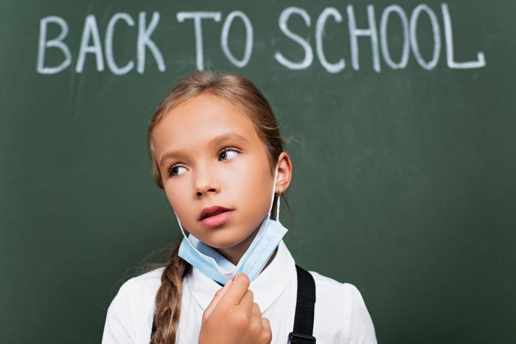 selective focus of exhausted schoolgirl taking off protective mask near chalkboard with back to school lettering - Photo, Image