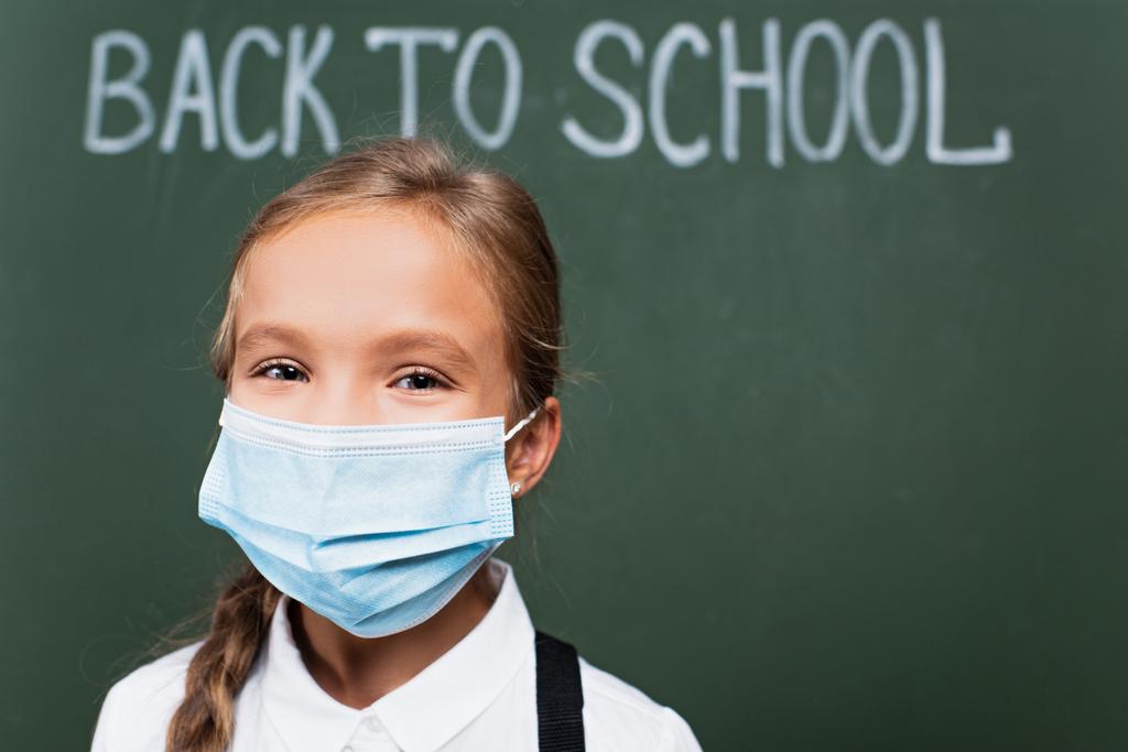 селективный фокус позитивной школьницы в медицинской маске, смотрящей на камеру рядом с доской с надписью   - Фото, изображение
