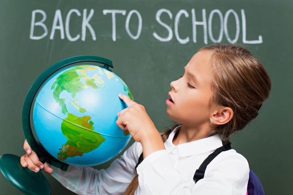 selective focus of attentive schoolgirl pointing with finger at globe near back to school lettering on chalkboard - Photo, Image
