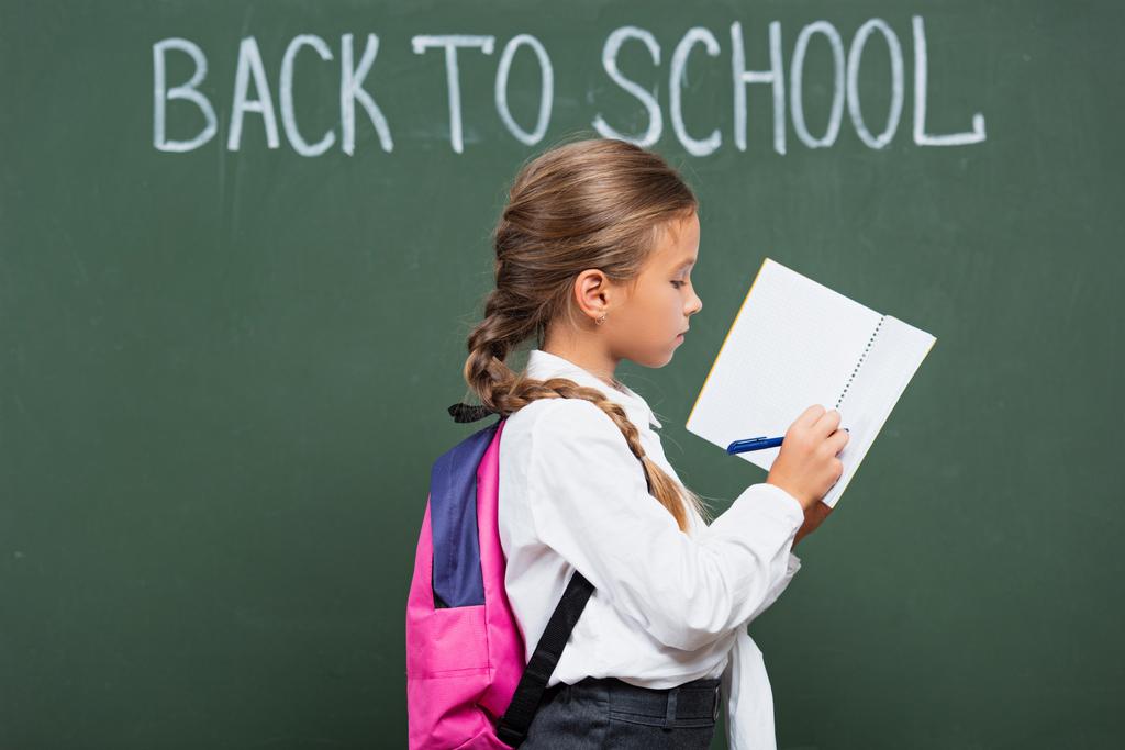 Sırt çantalı okul kızı, yazı tahtasının yanında yazı yazıyor, okula geri dönüyor. - Fotoğraf, Görsel