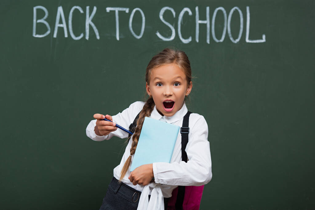 Aufgeregtes Schulmädchen zeigt mit Stift auf Buch neben Tafel mit Schulbuchstaben - Foto, Bild