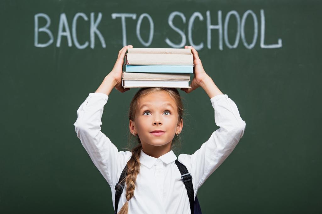 positive schoolgirl holding stack of books above head near chalkboard with back to school lettering - Photo, Image