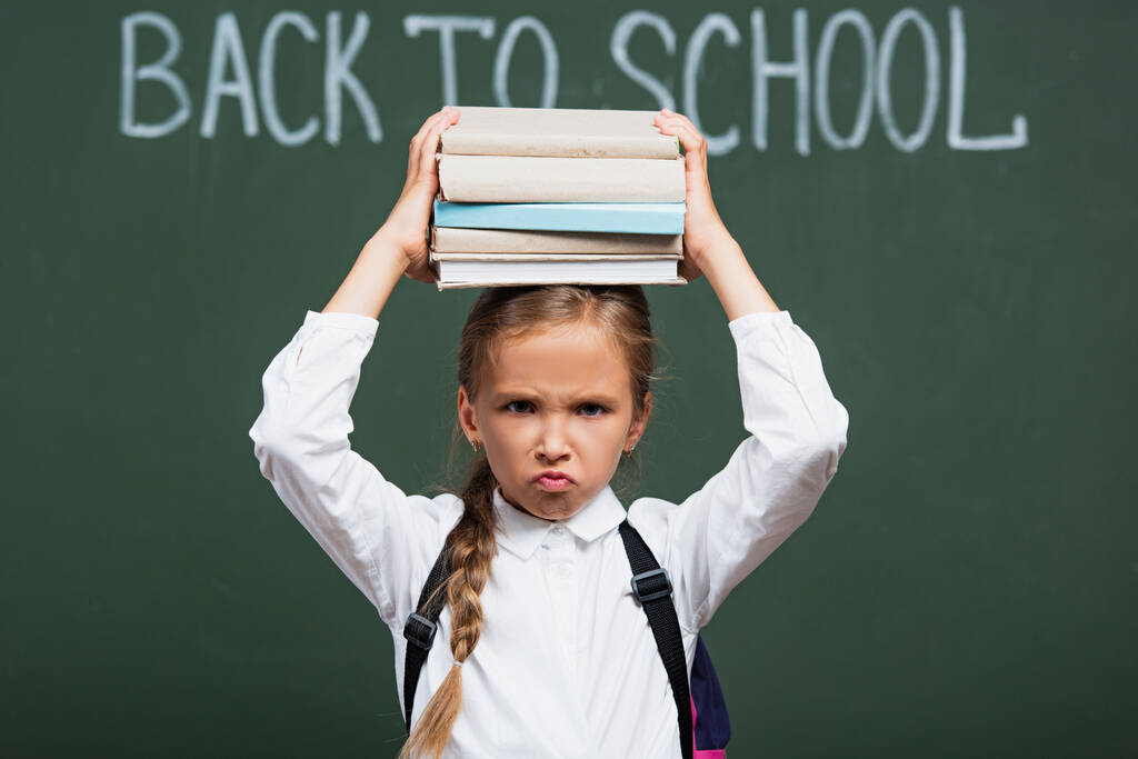 selective focus of displeased schoolgirl holding stack of books above head near chalkboard with back to school lettering - Photo, Image