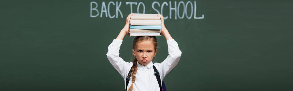 panoramic crop of displeased schoolgirl holding stack of books above head near chalkboard with back to school lettering - Photo, Image
