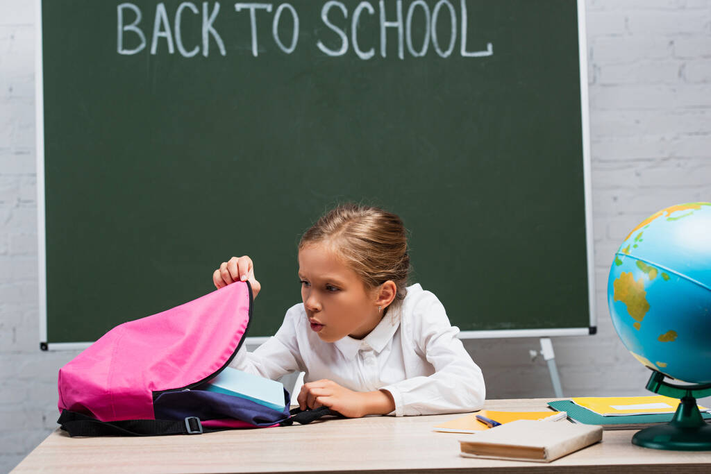 verrast schoolmeisje kijken in rugzak terwijl zitten aan een bureau in de buurt van globe en schoolbord met terug naar school belettering - Foto, afbeelding