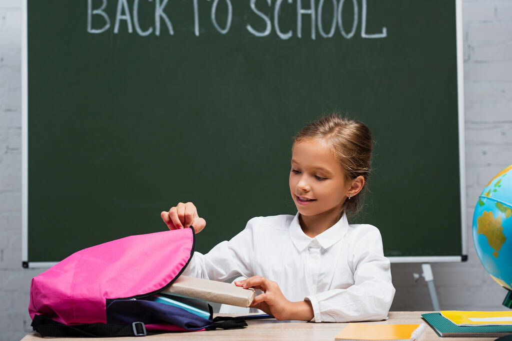 smiling schoolgirl taking books from backpack while sitting at desk near chalkboard with back to school inscription - Photo, Image