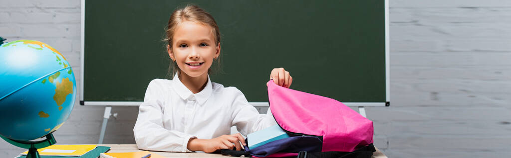 usmívající se školačka brát knihy z batohu, zatímco sedí za stolem poblíž zeměkoule a tabule - Fotografie, Obrázek