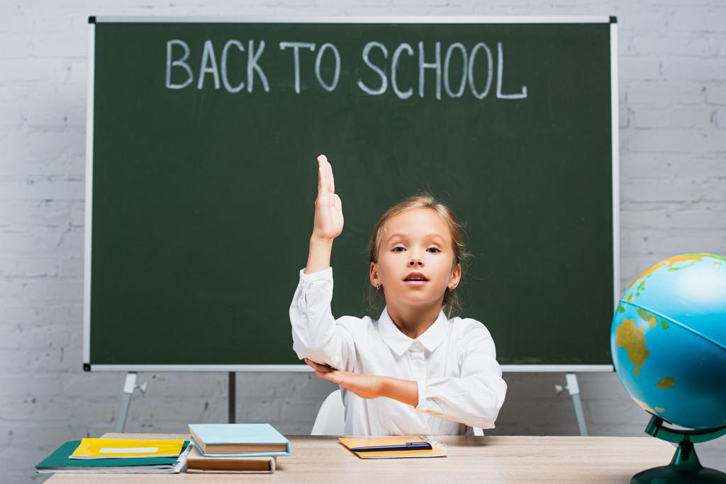 adorable schoolgirl raising hand while sitting at desk near globe and chalkboard with back to school inscription - Photo, Image