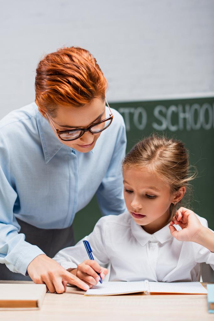 Внимательный учитель указывает пальцем на симпатичную школьницу, пишущую в книжке - Фото, изображение