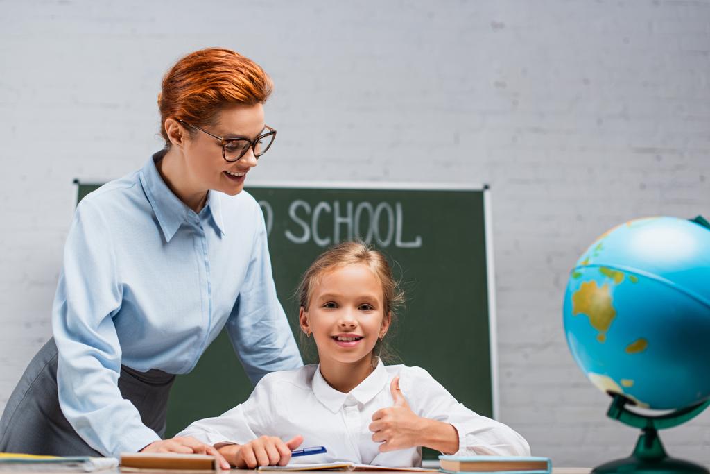 insegnante sorridente in piedi vicino a scolara felice seduto alla scrivania e mostrando pollice in su  - Foto, immagini