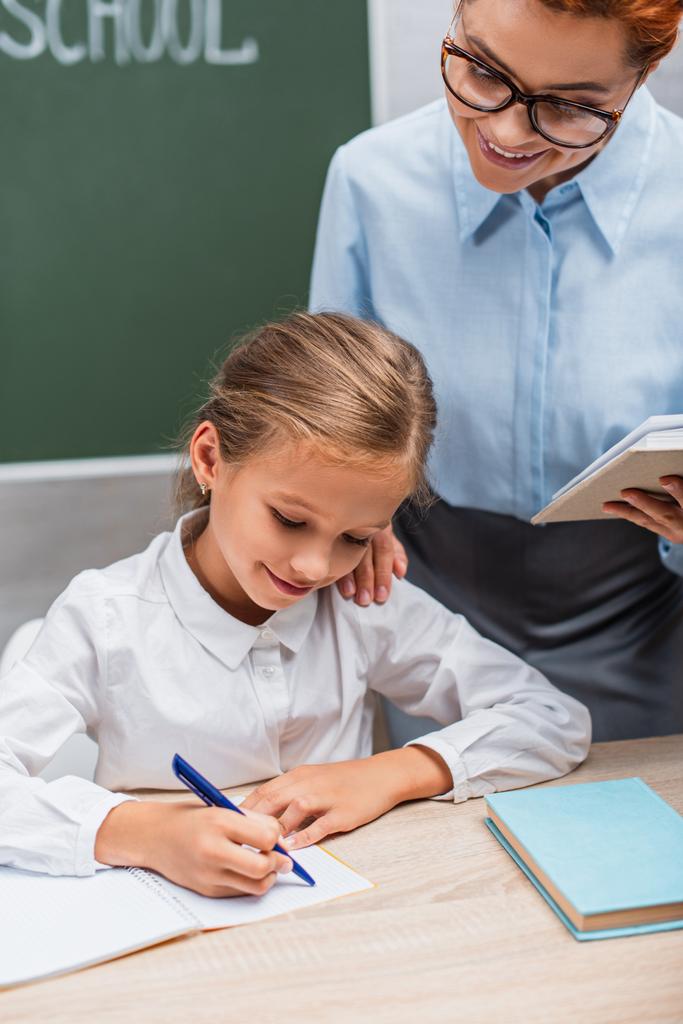 enseignant souriant regardant écolière assise au bureau et écrivant dans un cahier  - Photo, image