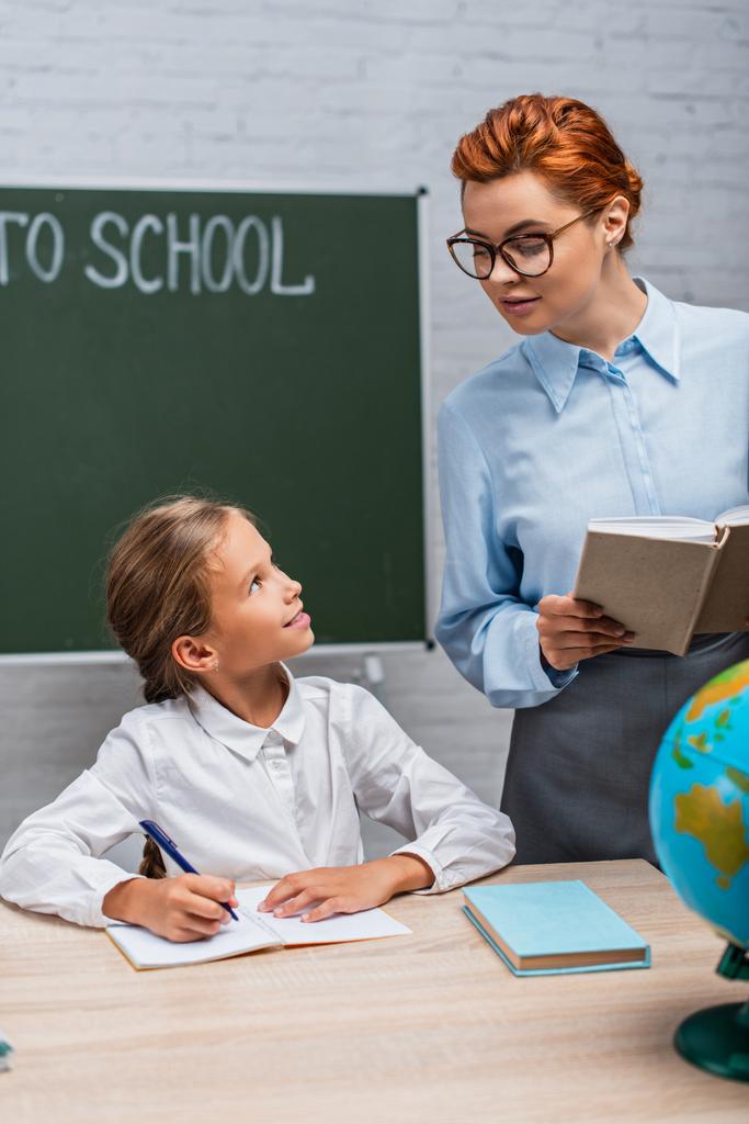 улыбающийся учитель смотрит на школьницу, сидящую за столом и пишущую в блокноте  - Фото, изображение