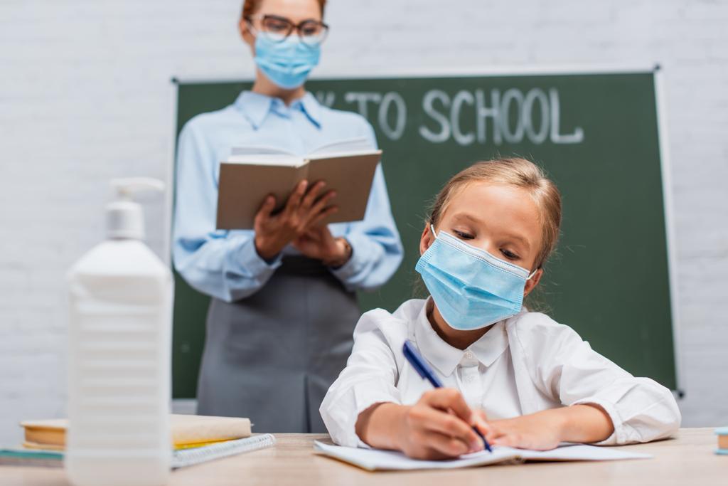 селективный фокус школьницы в медицинской маске, пишущей в блокноте, и учителя, стоящего рядом с доской с книгой - Фото, изображение