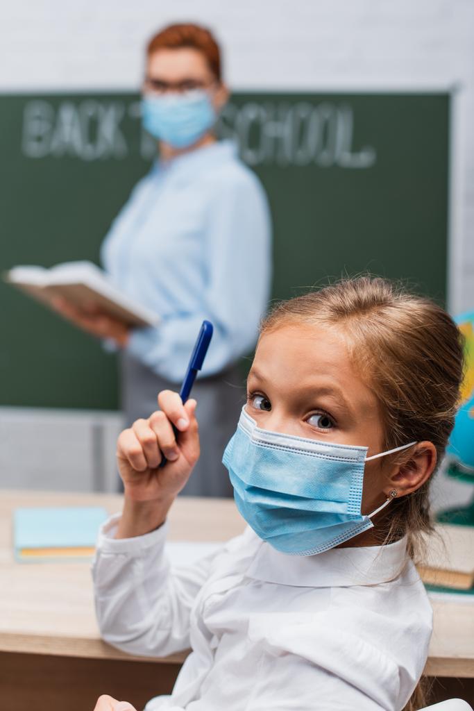 selective focus of schoolgirl in medical mask holding pen and looking at camera, and teacher with book near chalkboard - Photo, Image