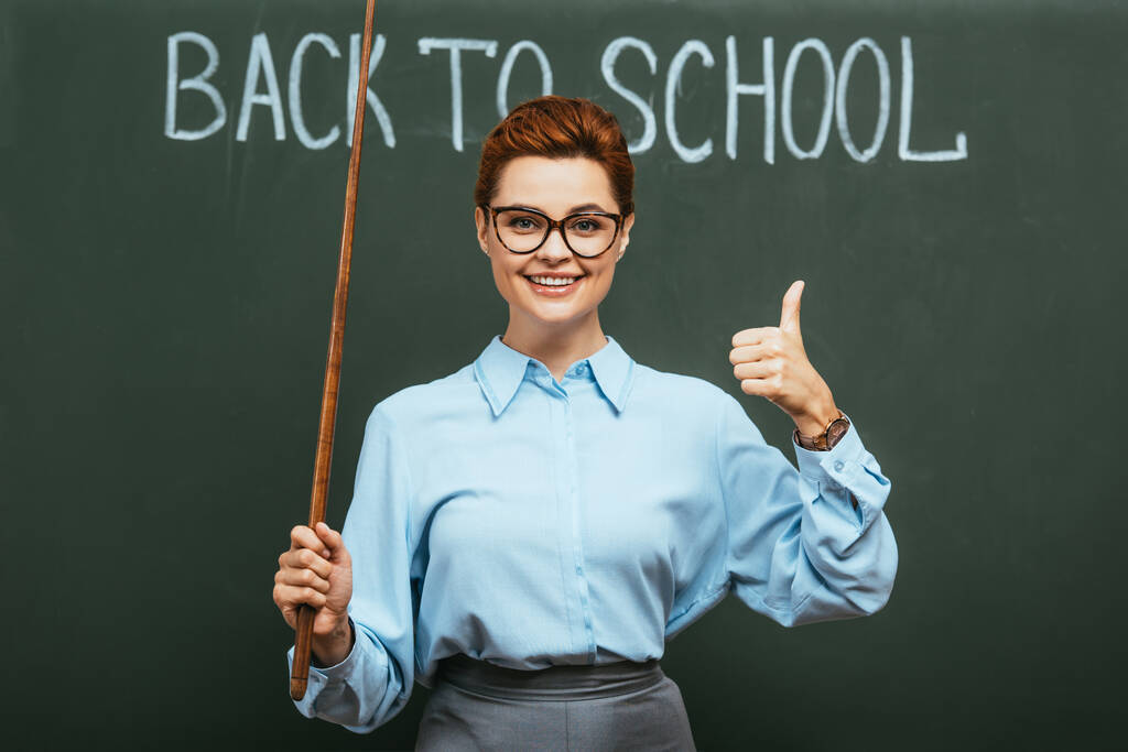 insegnante sorridente con bastone indicante mostrando pollice verso l'alto vicino lavagna con di nuovo a scuola lettering   - Foto, immagini
