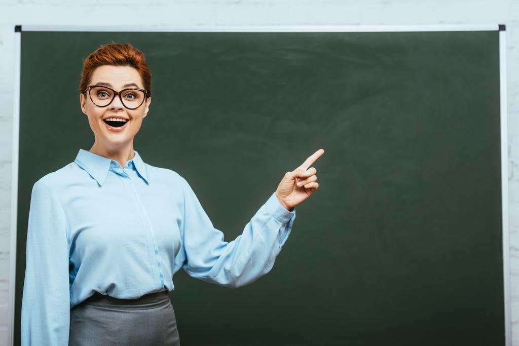 excited teacher looking at camera and pointing with finger near chalkboard  - Photo, Image