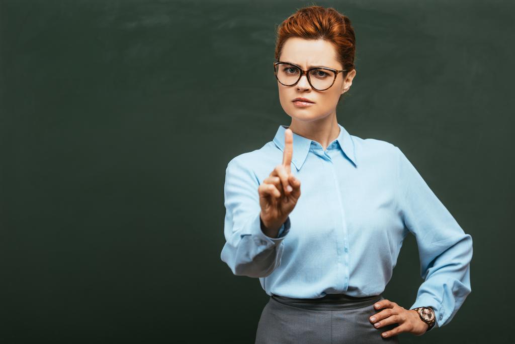 суворий вчитель в окулярах показує стоп- жест, стоячи біля крейдяної дошки
 - Фото, зображення