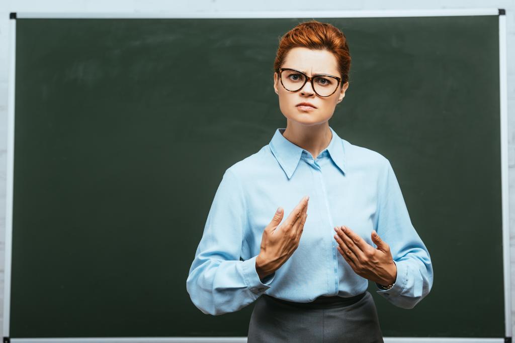 serious teacher in eyeglasses looking at camera while standing near chalkboard   - Photo, Image