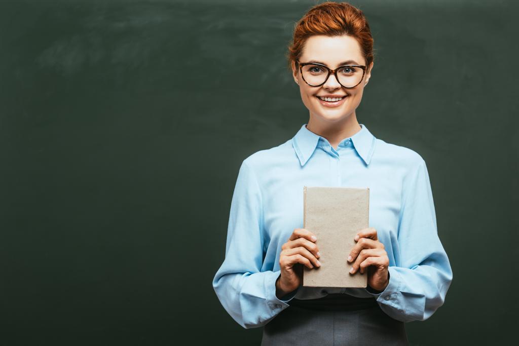 χαρούμενος δάσκαλος σε γυαλιά κρατώντας βιβλίο, ενώ στέκεται κοντά σε μαυροπίνακα   - Φωτογραφία, εικόνα