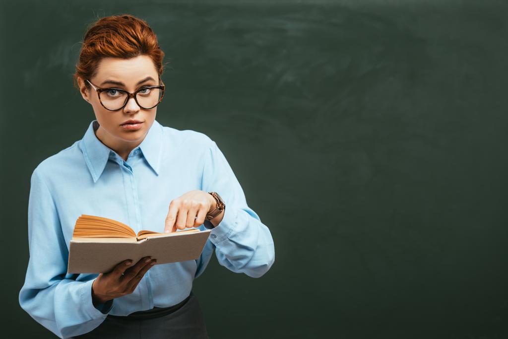 tarkkaavainen opettaja silmälaseissa osoittaa avoimeen kirjaan seistessään liitutaulun lähellä   - Valokuva, kuva