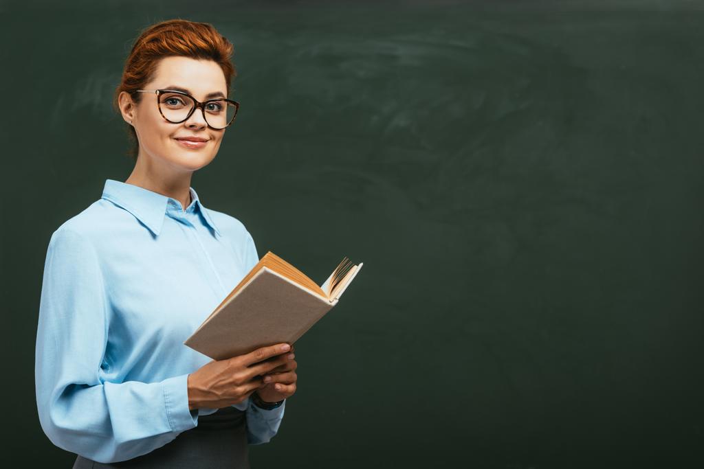 happy, beautiful teacher in eyeglasses holding open book while standing near chalkboard - Photo, Image