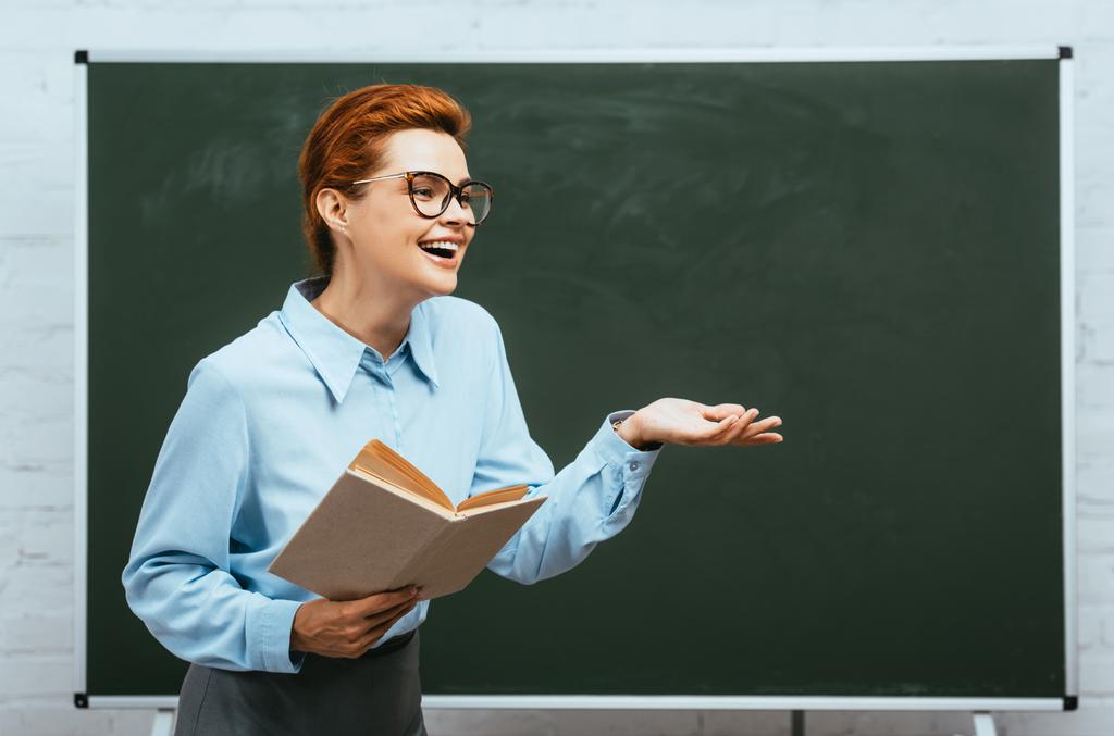 smějící se učitel v brýlích drží knihu, zatímco stojí u tabule s otevřenou paží - Fotografie, Obrázek
