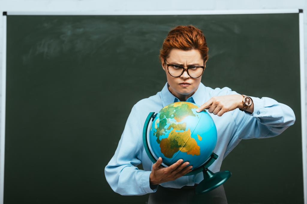 Strenger Lehrer zeigt mit dem Finger auf Globus, während er neben Tafel steht - Foto, Bild