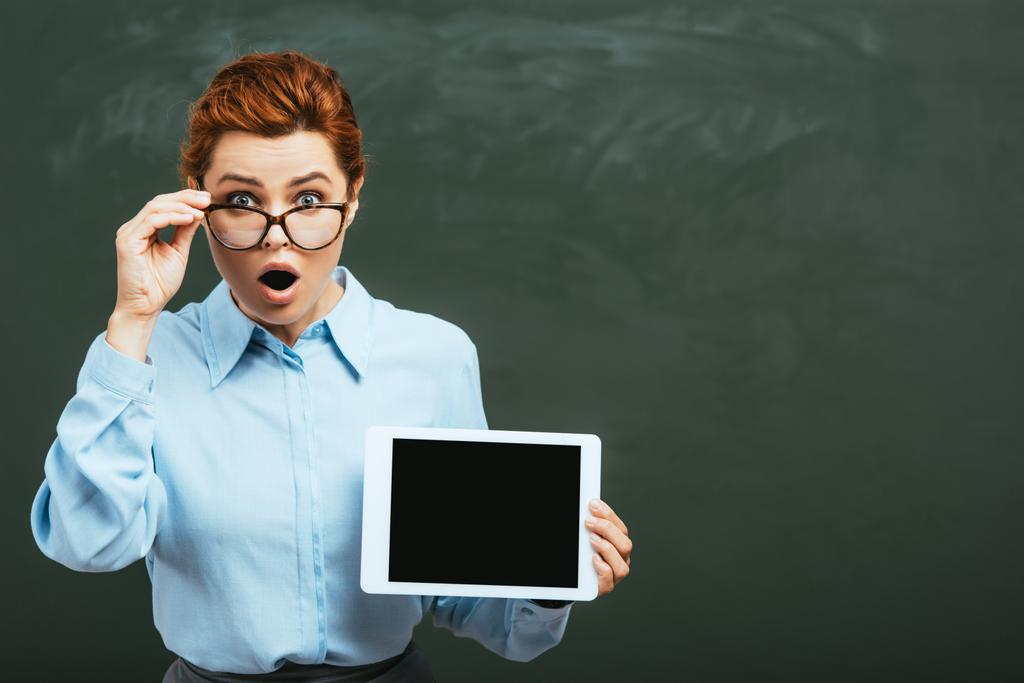 Schockierter Lehrer berührt Brille, während er digitales Tablet mit leerem Bildschirm in der Nähe der Tafel hält - Foto, Bild