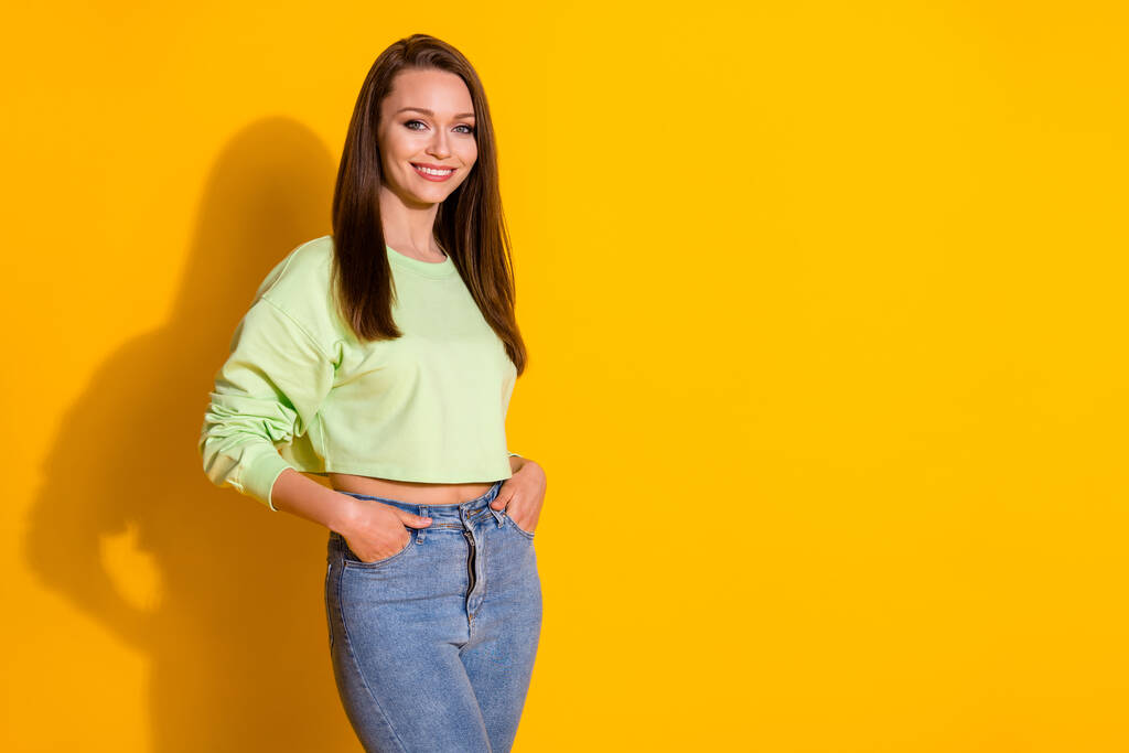 Фото довольно очаровательной молодой леди хорошее настроение зубастый улыбка носить зеленый свитер обнаженной брюшной полости джинсы изолированы яркий яркий ярко-желтый цвет фона - Фото, изображение