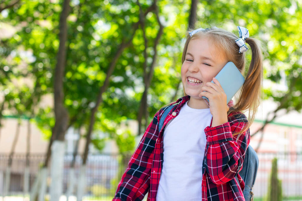 счастливая школьница с рюкзаком разговаривает по телефону на улице. в белой футболке и клетчатой рубашке - Фото, изображение