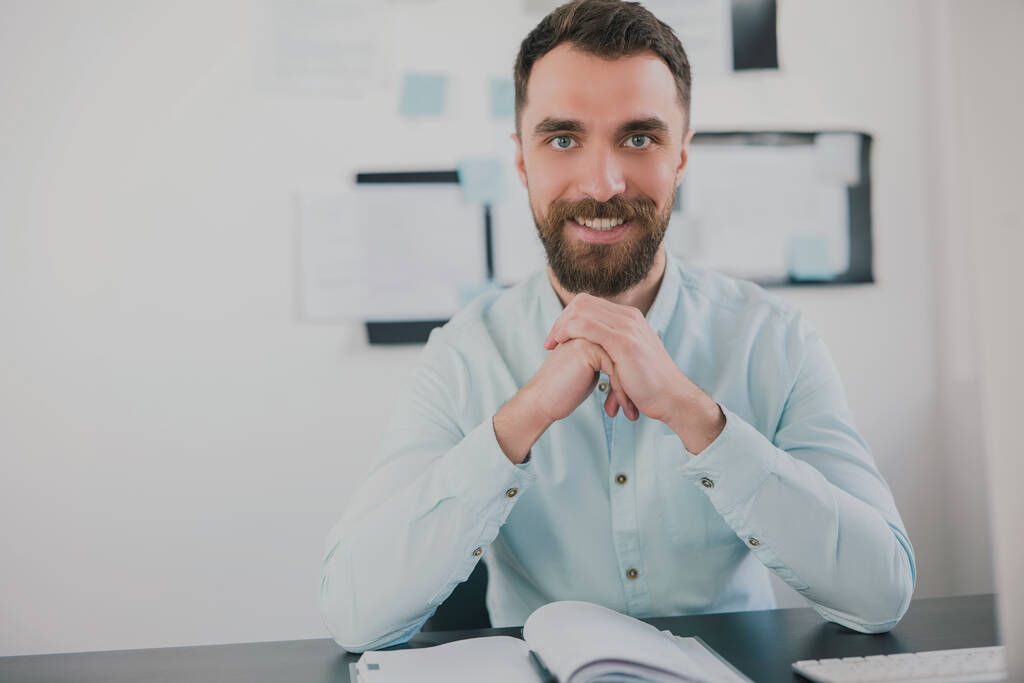 młody brodaty brunetka uśmiechnięty mężczyzna wygląda szczęśliwy podczas pracy nad projektem biznesowym w jego nowoczesnym biurze, trzyma się za ręce przy twarzy, rutynowa koncepcja pracy. - Zdjęcie, obraz