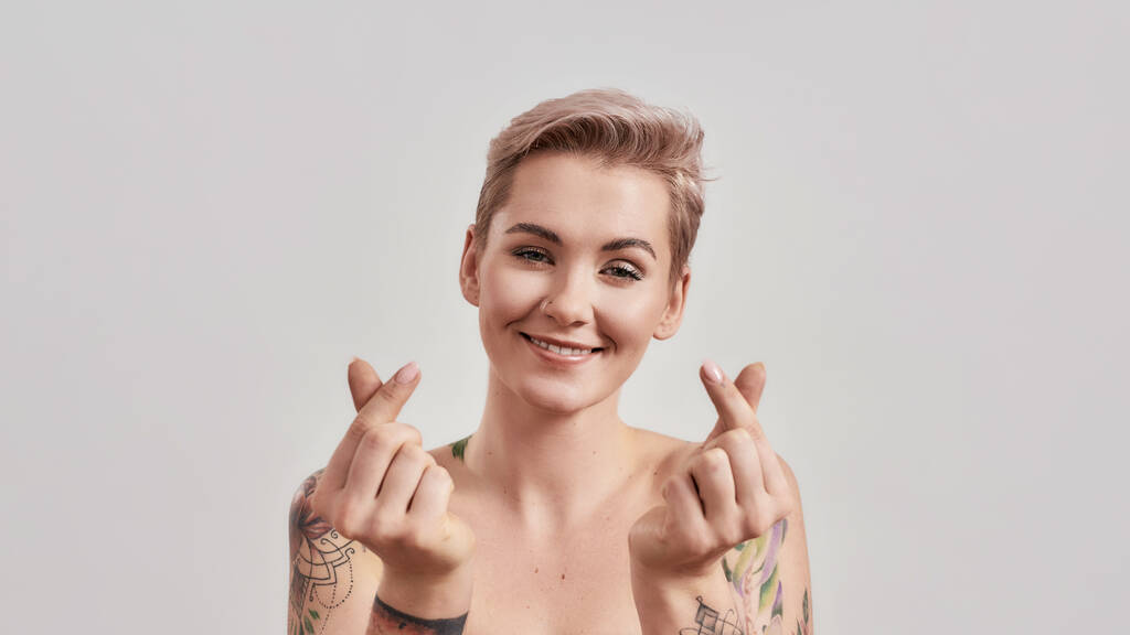 Spread Love. Portrait of cheerful half naked tattooed woman with pierced nose and short hair smiling at camera, showing Korean heart sign isolated over light background - Photo, Image