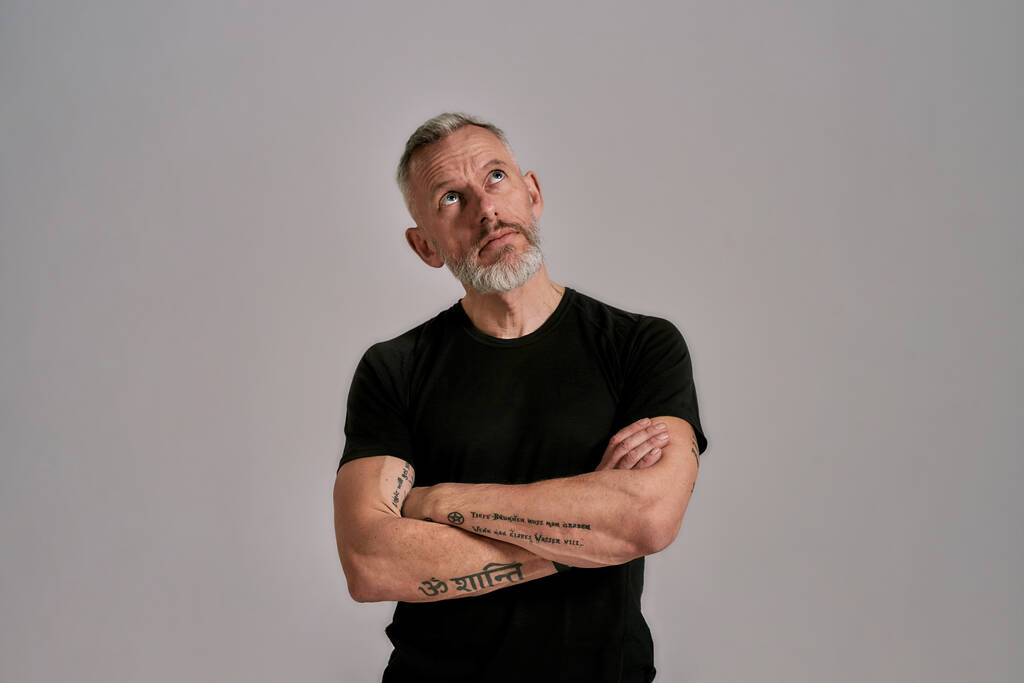 Homme d'âge moyen avec les bras croisés en t-shirt noir regardant de côté tout en posant en studio sur fond gris - Photo, image