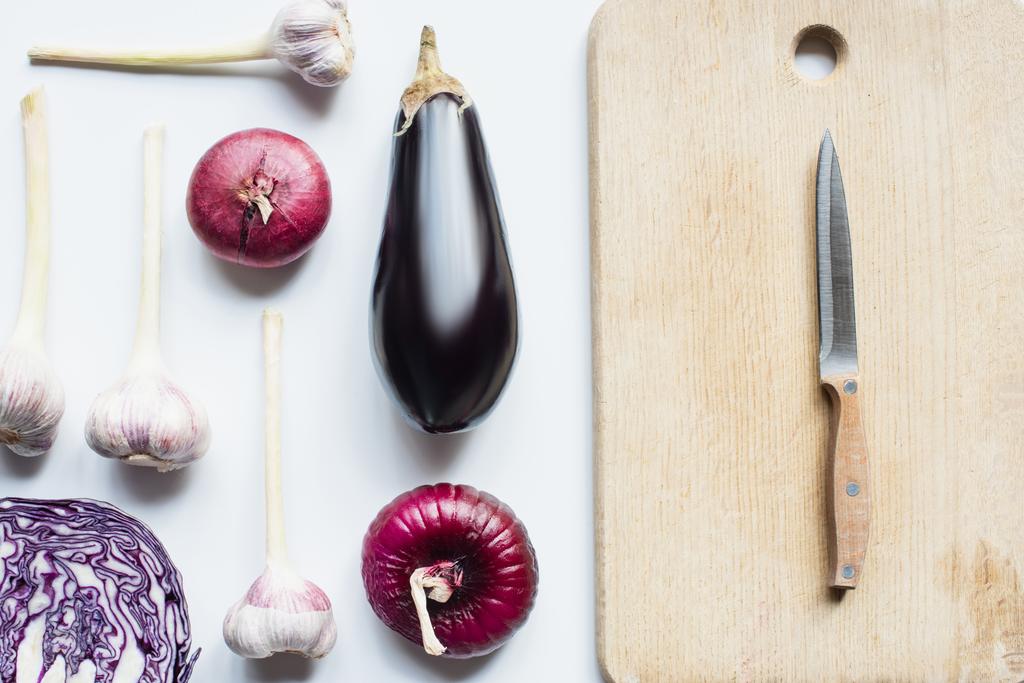 plat avec oignon rouge, chou rouge, aubergine et ail près de la planche à découper en bois avec couteau sur fond blanc - Photo, image
