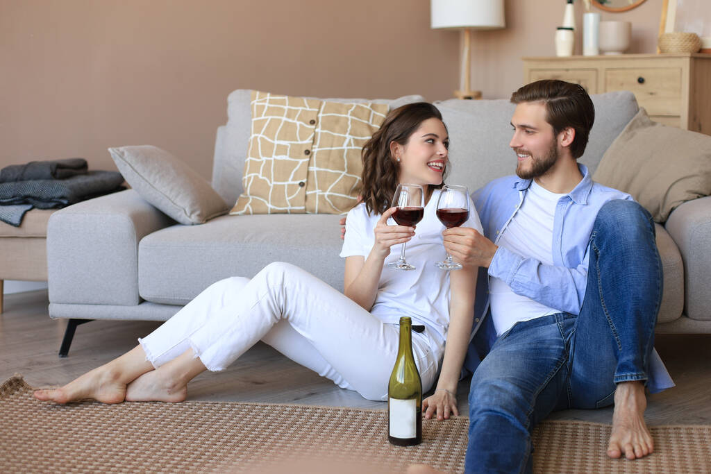 Szczęśliwa para siedzi, relaksuje się na podłodze w salonie, pije czerwone wino. Uśmiechnięty młody mąż i żona odpoczynku w domu cieszyć się romantyczną randkę na rodzinny weekend razem. - Zdjęcie, obraz