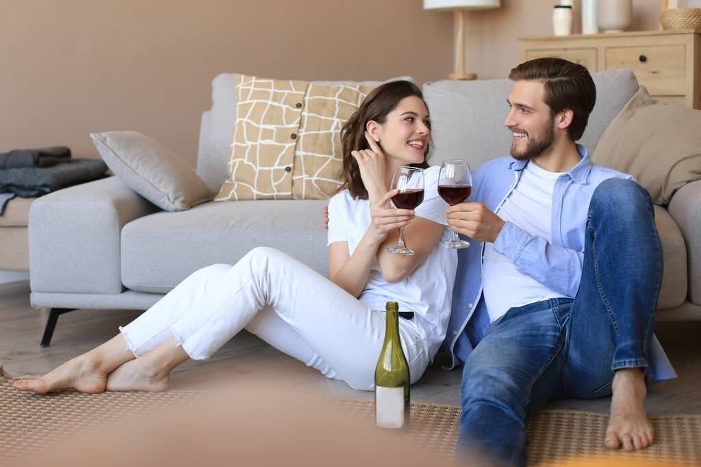 Ευτυχισμένο ζευγάρι κάθεται, χαλαρώνει στο πάτωμα στο σαλόνι, πίνοντας κόκκινο κρασί. Χαμογελαστοί νεαροί σύζυγοι ξεκουραστείτε στο σπίτι απολαύστε ρομαντικό ραντεβού το οικογενειακό Σαββατοκύριακο μαζί.. - Φωτογραφία, εικόνα