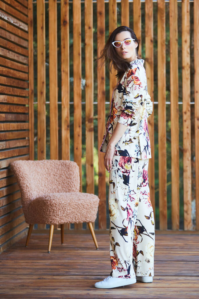 Jonge stijlvolle sexy vrouw in pyjama staande op terras in tropisch hotel, palmbomen achtergrond, lang zwart haar, zonnebril, oorbellen, zonnebril. - Foto, afbeelding