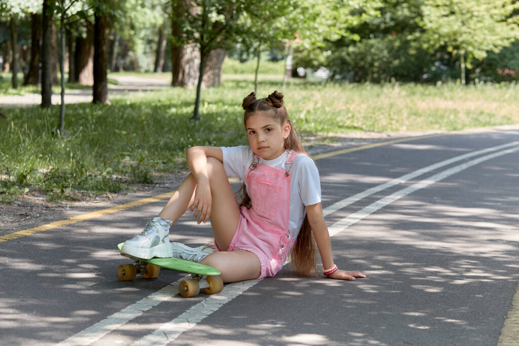 Όμορφη κοπέλα με μακριά σκούρα ίσια μαλλιά κάθεται στην πίστα με ένα skateboard. - Φωτογραφία, εικόνα