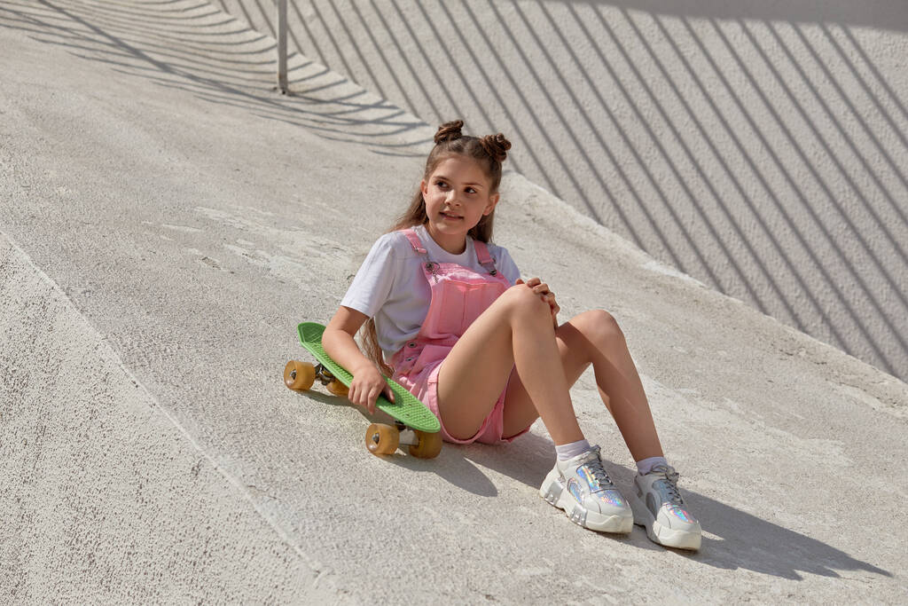 Uma menina bonita brilhante em um macacão na moda com um skate olha para o lado
 - Foto, Imagem