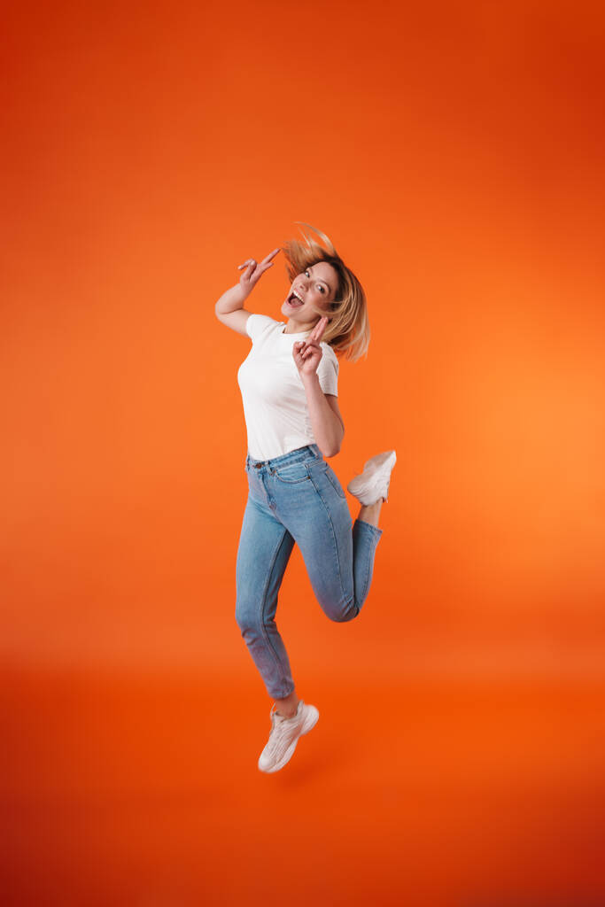 Образ веселой привлекательной женщины, демонстрирующей знак мира во время прыжка на оранжевом фоне - Фото, изображение