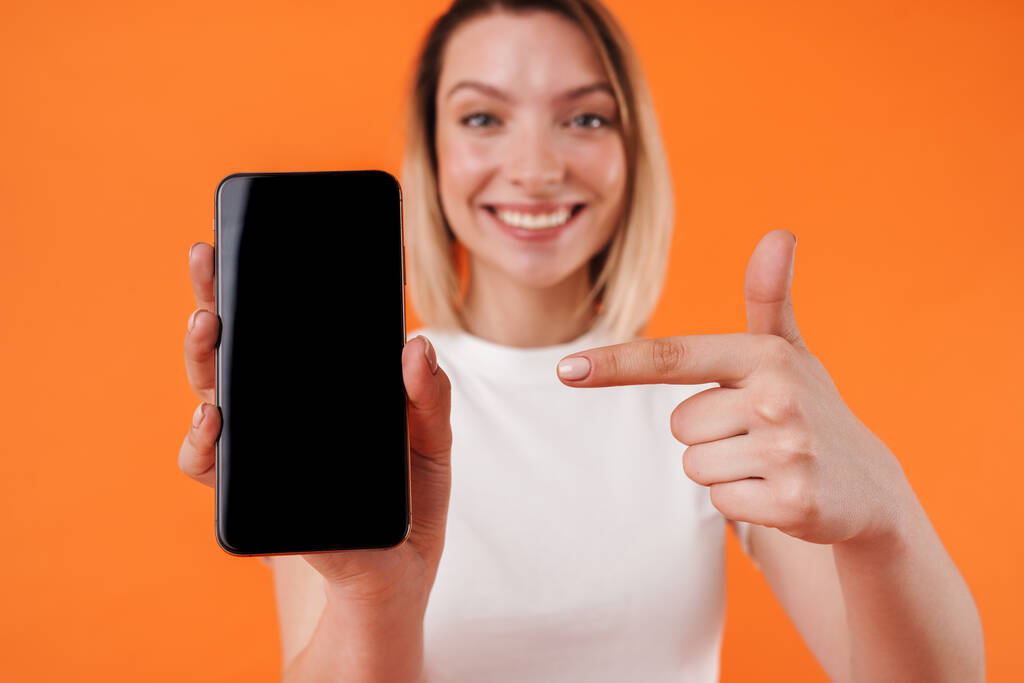 Kép vidám nő bemutató és ujjal mutogató mobiltelefon izolált narancssárga háttér - Fotó, kép