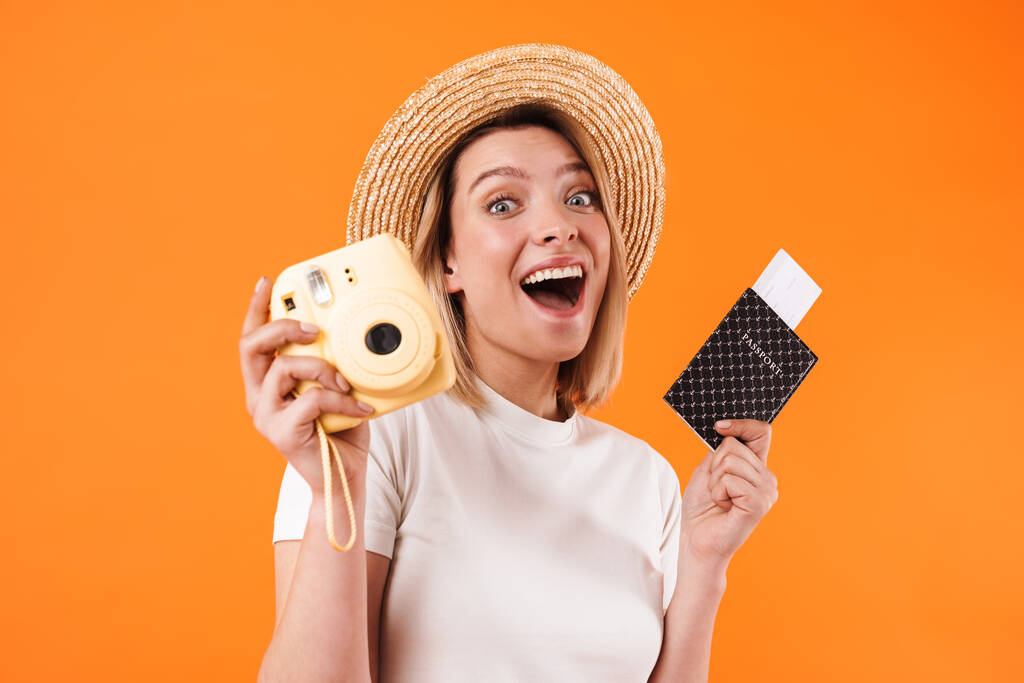 Imagen de una mujer sonriente con sombrero de paja sosteniendo pasaporte con pasajes y cámara aislada sobre fondo naranja
 - Foto, Imagen