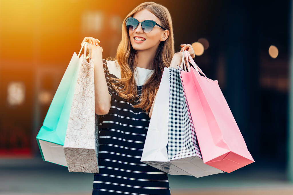 счастливая, красивая молодая женщина в солнечных очках и разноцветные сумки для покупок делает покупки на улице перед торговым центром. Магазины, мода - Фото, изображение