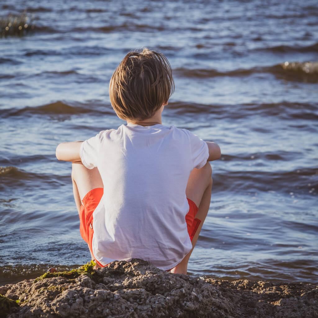 enfant assis sur le bord de la mer, garçon regarde dans la mer au coucher du soleil, concept de vacances d'été et de détente - Photo, image
