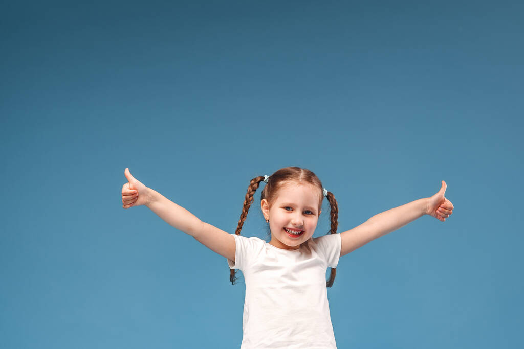petite fille avec des nattes dans un t-shirt blanc montre un geste d'approbation à la classe avec les deux mains et est heureux. Isolé sur un fond bleu. - Photo, image