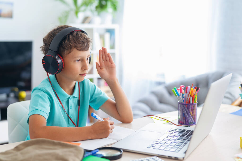 focused schoolboy boy wearing headphones uses laptop for distance learning during quarantine period. child raises his hand to answer lesson. e-education. Distance Learning, Home Learning. Stay at home - Photo, Image